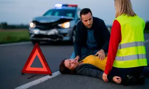 Un hombre y una mujer asisten a una persona en el suelo tras un accidente. Un triángulo de señalización y un coche dañado en el fondo ilustran la importancia de la seguridad vial.