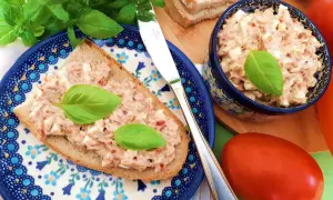 Pyszna pasta jajeczna z tuńczykiem na kromce chleba, ozdobiona bazylią. Obok miseczka z pastą i świeże pomidory.