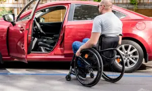 Uomo in carrozzina accanto a un'auto rossa, con portiera aperta. Un esempio di accessibilità, come previsto dall'art. 30 comma 7 legge 388 del 2000.