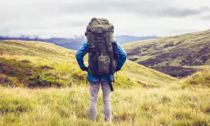 Wanderer mit großem Rucksack steht auf einem Hügel, bereit für seine 1 Jahr Weltreise. Die Landschaft ist grün und hügelig.