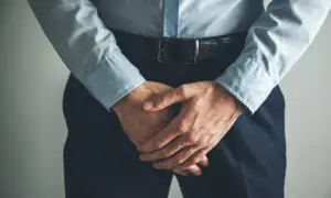 Un homme en chemise bleue et pantalon sombre, mains croisées devant lui, semble avoir un testicule qui gratte.