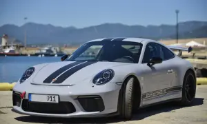 Szary Porsche Carrera 911 z czarnymi pasami na masce i dachu, zaparkowany przy nabrzeżu z widokiem na góry.