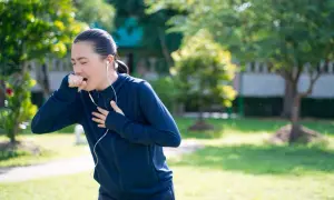 Donna con cuffie che corre in un parco, colpita da un attacco di tosse, si porta una mano alla gola.