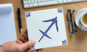 Dłoń trzyma kwadratową płytkę z symbolem Strzelca, znakiem zodiaku dla osób urodzonych 28 listopada.