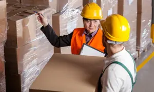 Kobieta w kasku i kamizelce omawia z pracownikiem normy dźwigania dla młodocianych, wskazując na paczki w magazynie.