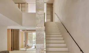 Escaleras de cemento blancas y minimalistas ascienden junto a un pilar de piedra texturizada, conectando niveles en un interior luminoso.