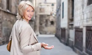 Uśmiechnięta kobieta z blond włosami w modnych, odmładzających krótkich fryzurach bob, zaprasza gestem ręki do wąskiej uliczki.