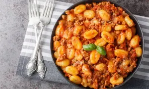 Un plat de gnocchi maison savoureux, nappés d'une sauce bolognaise riche et garnis d'une feuille de basilic frais. Deux fourchettes argentées sont posées sur une serviette à côté.