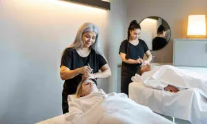 Dos terapeutas aplican tratamientos faciales a dos clientes en camillas. Una mujer con cabello plateado realiza un **olimpia masaje** facial a una cliente rubia.