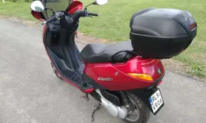 Czerwony skuter Peugeot Elyseo 125 z czarnym kufrem na bagaż, gotowy na podróż po malowniczych drogach.