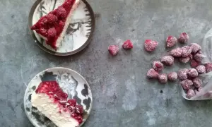 Dwa kawałki kremowego sernika z malinami na talerzach, obok garść mrożonych malin. Idealny deser na letnie popołudnie.