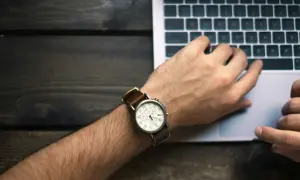 Nadgarstek z zegarkiem i dłonie na klawiaturze laptopa symbolizują nienormowany czas pracy.