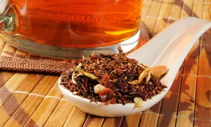 Suszone liście rooibos na łyżce, obok szklanki z bursztynowym naparem. Idealne do parzenia.
