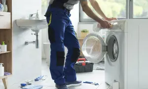 Instalator w niebieskich spodniach podłącza pralkę w łazience. Narzędzia na podłodze.