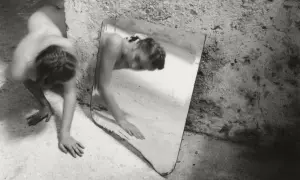 Une femme se contorsionne près d'un miroir brisé, son reflet fragmenté. Une œuvre de Francesca Woodman.