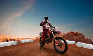 Motocyklista enduro w stroju i kasku na tle pustynnego krajobrazu z klifami i niebem.