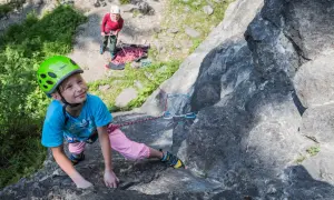 Ein Mädchen mit Helm klettert an einem Felsen, während eine andere Person sichert. Ein tolles Erlebnis beim **Klettern mit Kindern**.