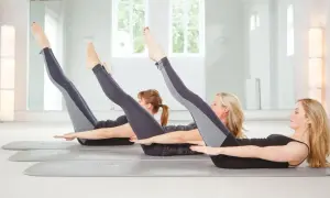 Tre donne eseguono posizioni pilates sul tappetino, sollevando gambe e braccia in un esercizio di core stability.