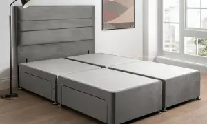 A grey sprung divan base with drawers, paired with a matching headboard, stands in a bright room with a large window.