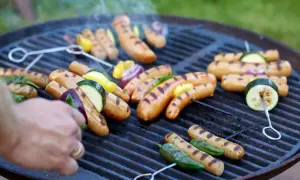 Grilla korv med grönsaker på en kolgrill. Röken stiger från de saftiga korvarna och grönsakerna som får fina grillränder.