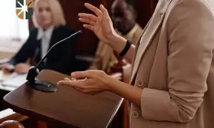 Oskarżyciel posiłkowy przemawia przy mównicy, gestykulując. W tle siedzą sędziowie.