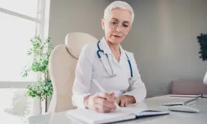 Lekarka w białym kitlu z niebieskim stetoskopem pisze w notatniku przy biurku.