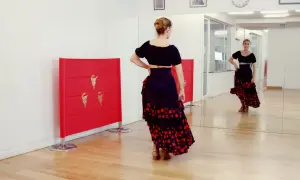 Bailarina con falda flamenca y top negro, practicando una rumbita flamenca frente a un espejo y paneles rojos con detalles dorados.
