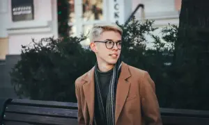 Młody mężczyzna w okularach i płaszczu prezentuje sportową elegancję, siedząc na ławce wśród zieleni.