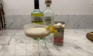 En krämig limoncello drink med citronskal, bredvid flaskor med limoncello, gin och vispgrädde.