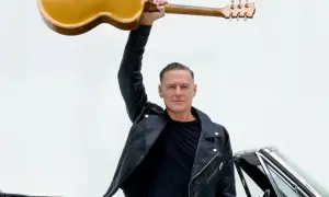 Bryan Adams w skórzanej kurtce unosi gitarę nad głową, gotowy na koncert. Obok klasyczny samochód.