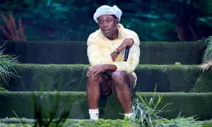 Igor, czyli Tyler, The Creator, siedzi na scenie w żółtej kurtce i jasnoniebieskiej czapce, trzymając mikrofon.