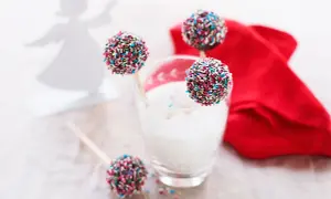 Färgglada cake pops med strössel står i ett glas med mjölk. En röd duk och en ängel i bakgrunden skapar en festlig stämning.