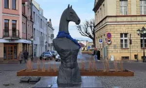 Pomnik z głową konia i niebieską szarfą, przypominający herb. W tle kamienice i ulica.