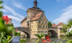 Das Alte Rathaus ragt über den Fluss in der Bamberger Innenstadt. Ein Fachwerkhaus mit gelben Balken und roten Ziegeln schmückt die Brücke.