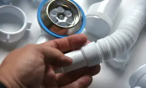 Dłoń trzyma element syfonu do wanny. Widać metalowy przelew z niebieską uszczelką i białe rury.