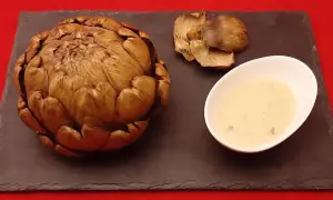 Un artichaut entier, cuit à la cocotte minute, servi avec sa sauce et quelques feuilles détachées sur une ardoise.