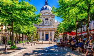 En solig paris april med gröna träd som ramar in en vacker byggnad. Folk sitter vid cafébord i skuggan.