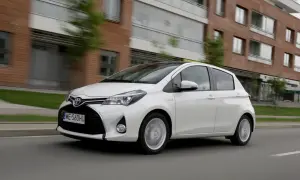 Biały model toyoty na 5 liter, Yaris Hybrid, jedzie ulicą obok nowoczesnych budynków.