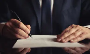 Mężczyzna w garniturze pisze długopisem na dokumencie. Dowiedz się, jak napisać testament bez notariusza.