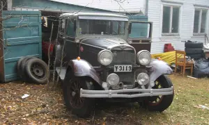 En gammal Dodge-bil står parkerad utanför ett garage. Bilen är mörkt vinröd med kromdetaljer och har en klassisk design.