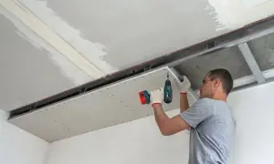 Mężczyzna montuje płyty regips do stelaża sufitowego, używając wkrętarki.