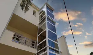 Moderno montacarichi persone esterno in vetro e metallo bianco, che sale lungo un edificio color crema, con cielo nuvoloso al tramonto.