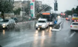 Gouttes de pluie sur un pare-brise, floutant la circulation urbaine. Les feux position seuls des voitures et des bus créent des traînées lumineuses sur la chaussée mouillée.