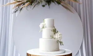 Eleganckie torty weselne wzory: biały, trzypiętrowy tort z delikatnymi kwiatami i koronkowym zdobieniem, prezentowany na złotym postumencie.
