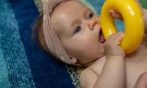 Niemowlę w opasce na głowie wkłada do buzi żółtą zabawkę na tle kolorowego koca.