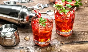 Zwei rote Cocktails mit Erdbeeren und Minze, Eiswürfel, Cocktailshaker und Barzubehör auf Holztisch.