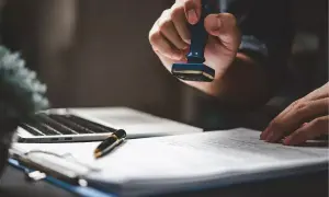 Dłoń z pieczęcią nad dokumentem, symbolizującym notarialne poświadczenie dziedziczenia. Obok laptop i długopis.