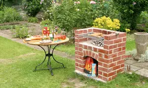 Ein selbstgebauter Ziegelgrill mit Würstchen und Kartoffeln, bereit für ein Gartenfest. Ein Tisch mit Salaten und Getränken steht daneben.