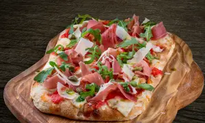 En avlång pinsa pizza med prosciutto, ruccola och parmesan på en träbricka.