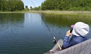 Osoba siedzi nad jeziorem Zagnanie, łowiąc ryby. W tle zielone drzewa i trzciny.